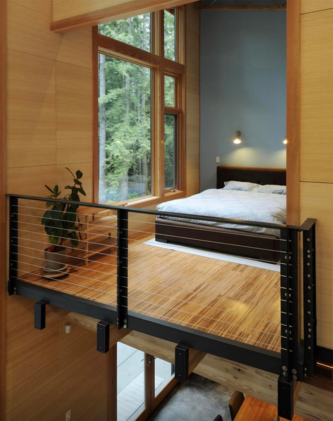 A Modern Two-Story Interior With A Loft Bedroom. The Loft Features A Bed With A Small Bedside Wall Lamp. A Large Window Provides A View Of Green Trees Outside. Below The Loft, There Is Open Space With Wood And Metal Design Elements, And Plants For Decoration.