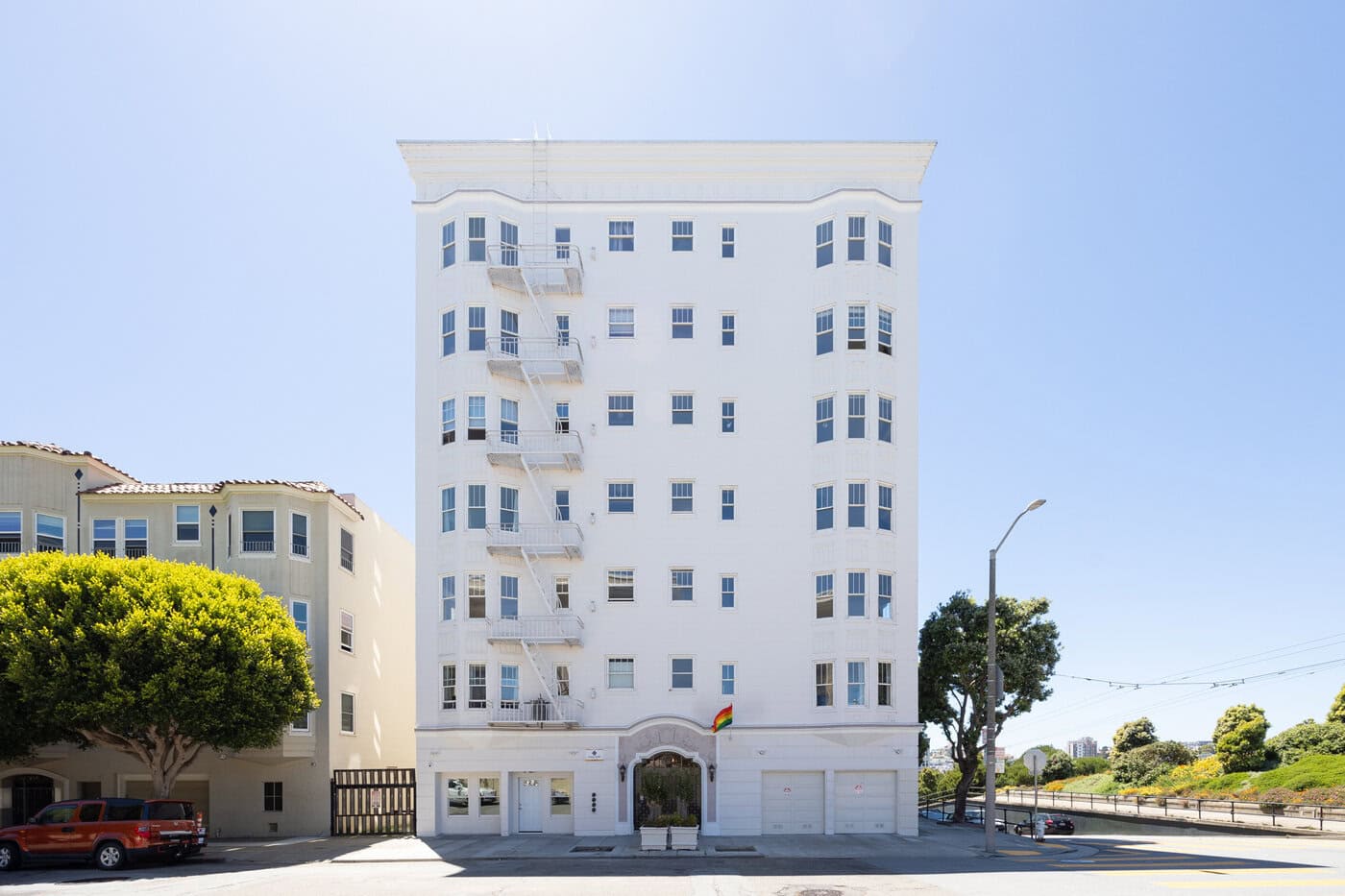 A Tall, White Apartment Building Stands Under A Clear Blue Sky. It Features Multiple Windows And A Fire Escape On The Front. A Few Trees And Parked Vehicles Are Visible Nearby On The Quiet Street. A Rainbow Flag Hangs Near The Entrance.