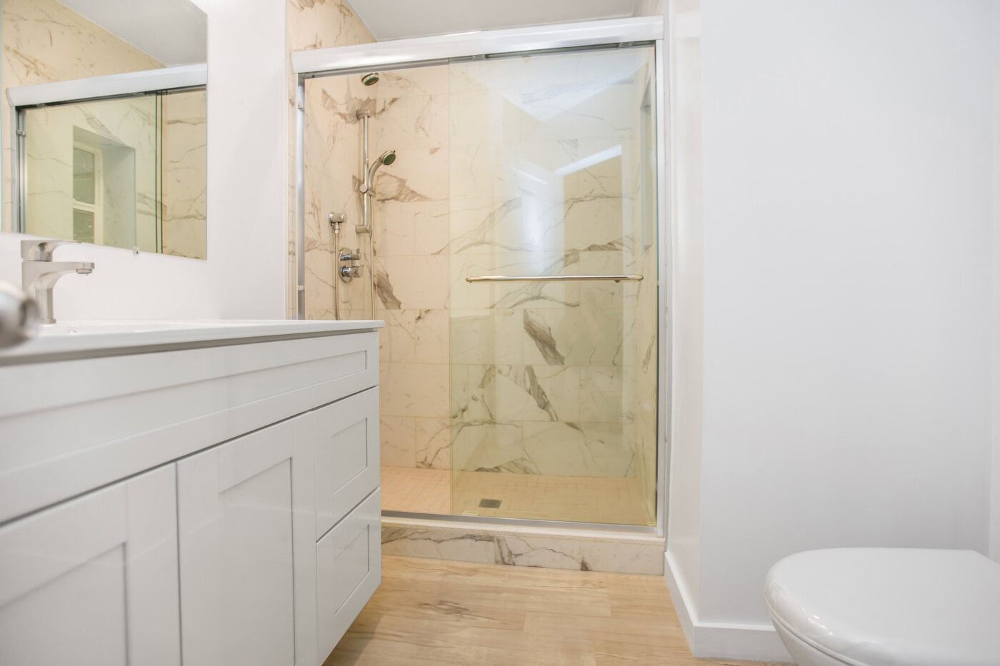 Bright Bathroom Featuring A Walk-In Shower With Glass Doors And Marble-Patterned Tiles. A White Vanity With A Sink Is On The Left, And A Toilet Is Visible On The Right. The Floor Is Light Wood, Enhancing The Clean And Modern Aesthetic.