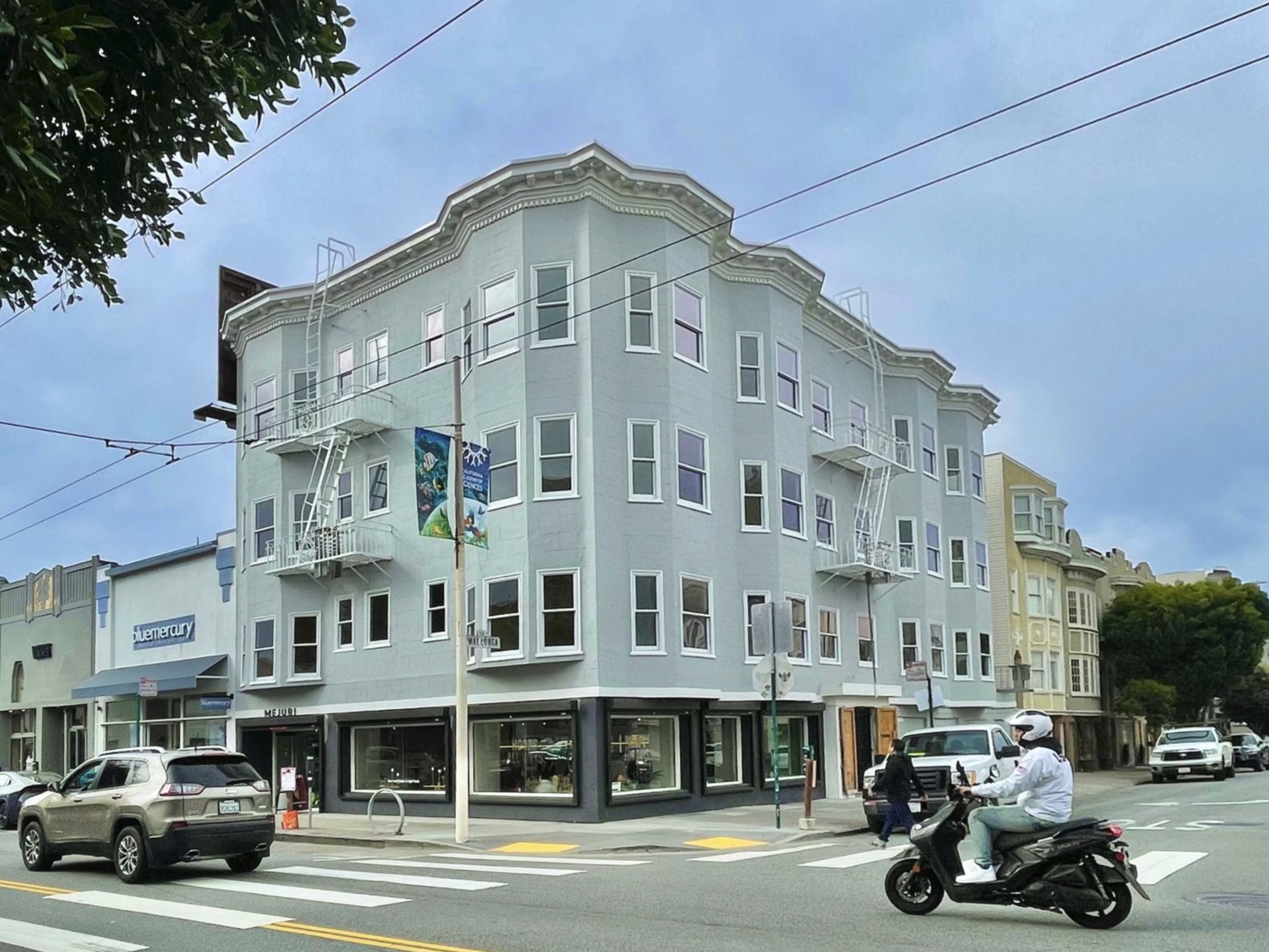 A Person On A Scooter And A Car Pass A Gray, Four-Story Corner Building With Shops At Street Level And Bay Windows Above, Under A Cloudy Sky At An Urban Intersection.