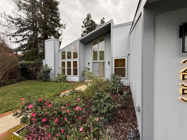 A Modern Gray House With Large Triangular Windows, A Sloped Roof, And A Landscaped Front Yard With Green Grass, Bushes, And Pink Flowers, Set Under An Overcast Sky.